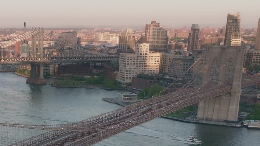 Aerial view shows bridges connecting parts of New York City. Buildings line the shore and the river reflects light from the setting sun. People move across the bridges.