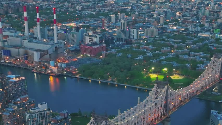 New York skyline shows buildings and parks by the water with lights. The Queensboro Bridge connects areas across the river. This scene captures city life at night.