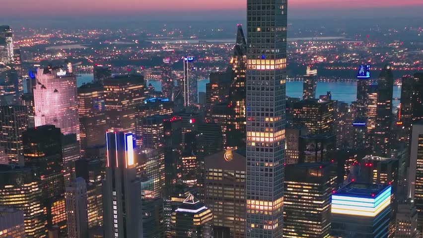 The scene shows the New York skyline during dusk with buildings lit up. The lights from the city shine bright. Rivers are visible in the distance as day turns to night.