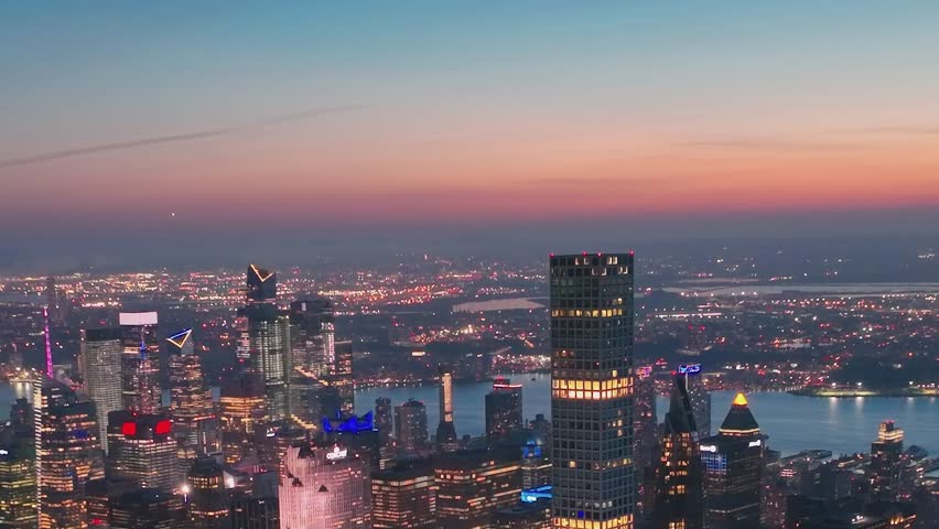 The scene shows the skyline of New York City during sunset with lights glowing from tall buildings. The sky changes color as day turns to night.