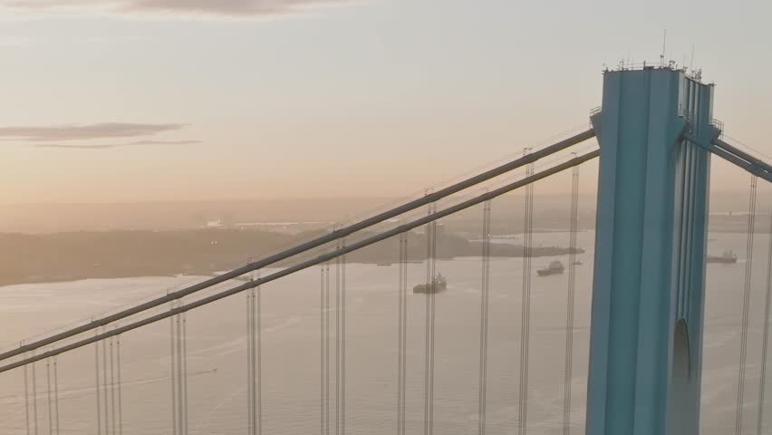 Aerial view shows a large bridge in New York. The sun sets behind the water, casting light on the bridge and boats below. The scene captures the busy city life.