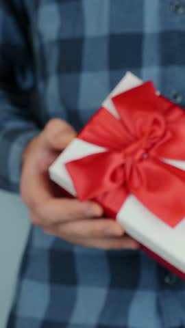 Man holds and shows white gift box with tied red bow, moves towards to camera, close up. Present for St. Valentine's Day in male hands. Vertical video