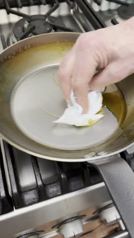 Vertical video - Closeup of a man’s hand lifting out some paper towel soaked with leftover cooking oil from a steel frying pan on a kitchen cooker hob.