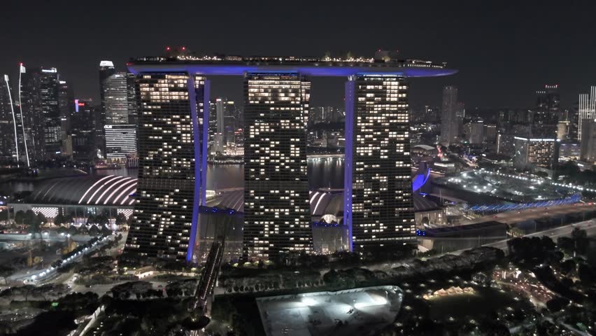Singapore – July 2024.
Aerial drone view of a major urban avenue in Singapore showing dense residential and commercial high-rise buildings bordering a wide multi-lane roadway with active traffic flow. The camera faces directly along the avenue, creating strong linear perspective as the road extends toward the distant skyline under an overcast sky. Cars, buses, and public transportation vehicles move in both directions, while pedestrian bridges and intersections connect surrounding districts. The footage documents daily urban activity, transportation infrastructure, and the metropolitan scale of Singapore.
