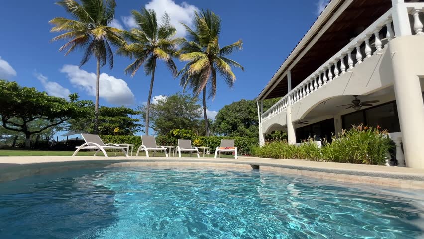 Low poolside view tracking shot at a luxury condo with waterfall feature overlooking palm trees blowing in the wind.