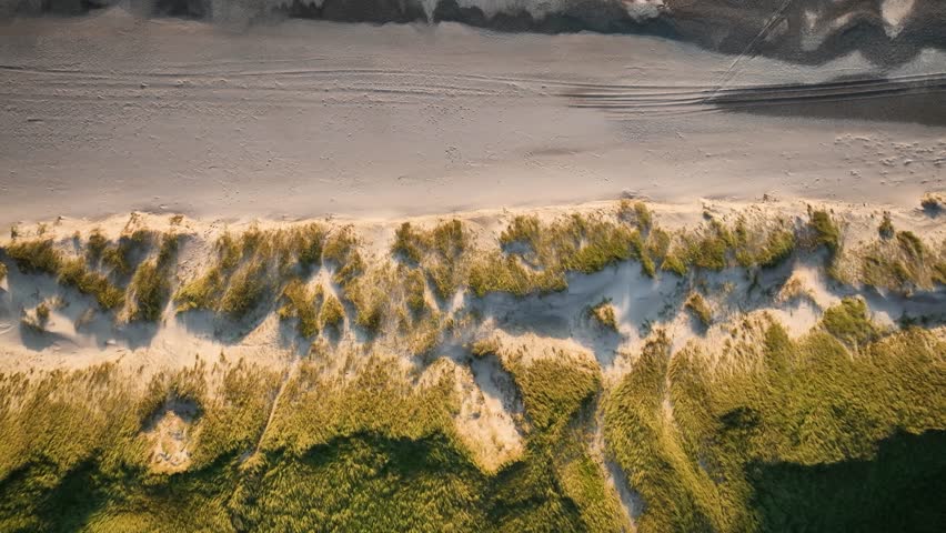 Aerial footage of a coastline with textured sand dunes covered in coastal grass, waves rolling toward shoreline and warm, shimmering sunset light.