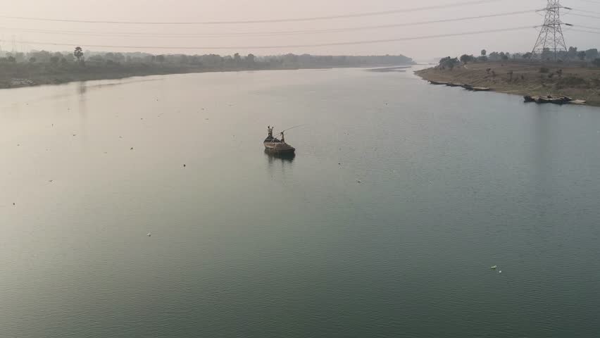 Two people are fishing in a small boat on a wide river in the distance. A small wooden boat with two people in it floats on a wide, calm river under a hazy, cloudy sky. The scene evokes a sense of peaceful and enchanting evening tranquility.