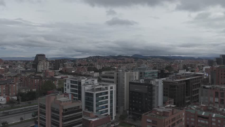 Bogotá, Bogotá D.C., Colombia – July 2024:
Aerial view of a major urban avenue in Bogotá, Colombia, showing dense residential and commercial buildings along a wide multi-lane roadway with moving traffic. The drone shot looks straight along the avenue, creating strong linear perspective as the road stretches toward the distant Andean mountains under an overcast sky. Vehicles, buses, and cars are visible traveling in both directions, while pedestrian bridges and intersections connect different parts of the city. This footage illustrates daily urban life, transportation infrastructure, and the scale of Colombia’s capital city.
