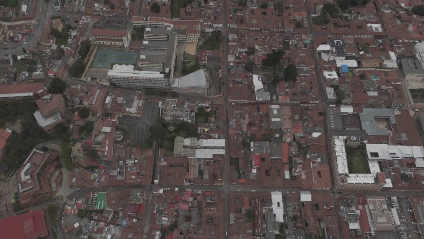 Bogota, Colombia; March 21, 2025:
Aerial drone footage showing a top-down and oblique view of downtown Bogota, the capital city of Colombia. The scene captures a dense historic urban area with tightly packed low-rise buildings, red tile rooftops, narrow streets, residential blocks, educational complexes, and mixed-use infrastructure.
The footage reveals the characteristic layout of Bogota’s historic center, where colonial-era architecture blends with modern buildings and institutional structures. Streets, courtyards, sports courts, trees, and public spaces are visible, illustrating everyday city life and urban density in one of South America’s largest capitals.
This aerial video documents architecture, city structure, and residential patterns in central Bogota, providing strong visual context for topics related to Latin American cities, urban development, population density, and metropolitan life. Suitable for editorial use in news media, documentaries, travel content, urban studies, and educational projects.
