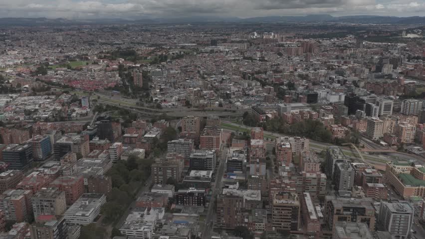 Bogotá, Bogotá D.C., Colombia – July 2024:
Aerial panoramic view of Bogotá, Colombia, showing a dense urban landscape with residential and commercial buildings intersected by major roads and highways. The drone footage highlights the city’s infrastructure, apartment blocks, office buildings, and green corridors integrated into the urban fabric. In the distance, the metropolitan area extends toward surrounding hills under a cloudy sky, reflecting Bogotá’s high-altitude geography and the scale of everyday life in Colombia’s capital city.