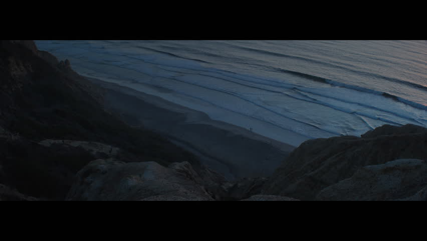 A sweeping dusk view of Black’s Beach from the steep Torrey Pines cliffs, capturing hikers making their way up the rugged trail while small groups of people stroll along the shoreline below. Soft blue
