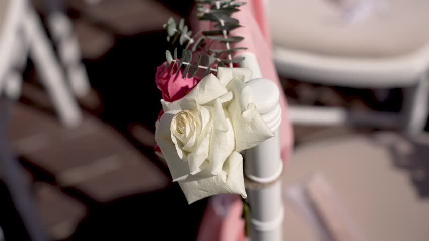 Fresh flowers are attached to white chairs in a garden. The roses add color and charm to the outdoor setup for a special gathering taking place in the daytime.