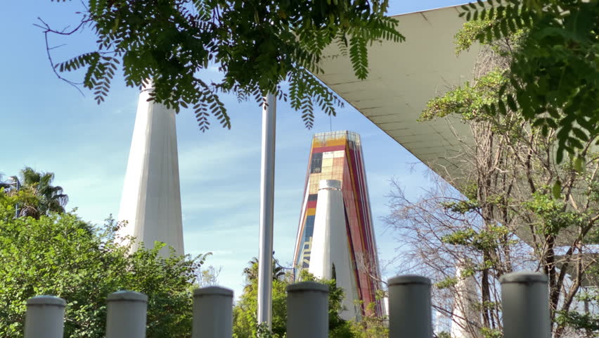 Expo 92 towers rise above dense greenery in Seville, white cones and a colorful central structure contrasting against a clear blue sky