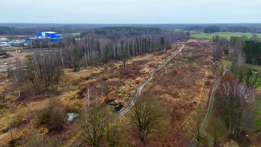 Aerial view of an overgrown access road cutting through mixed woodland and open terrain near a distant industrial complex on a cloudy late-autumn day.
