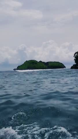 Wide shot of blue ocean waves with lush green tropical islands and a dramatic cloudy sky in the background, Indonesia