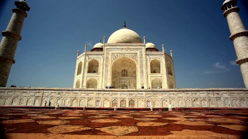 Iconic Taj Mahal in Agra, India rises against clear blue sky. Sunlight illuminates its stunning marble exterior. Visitors admire beautiful surroundings and historical significance.