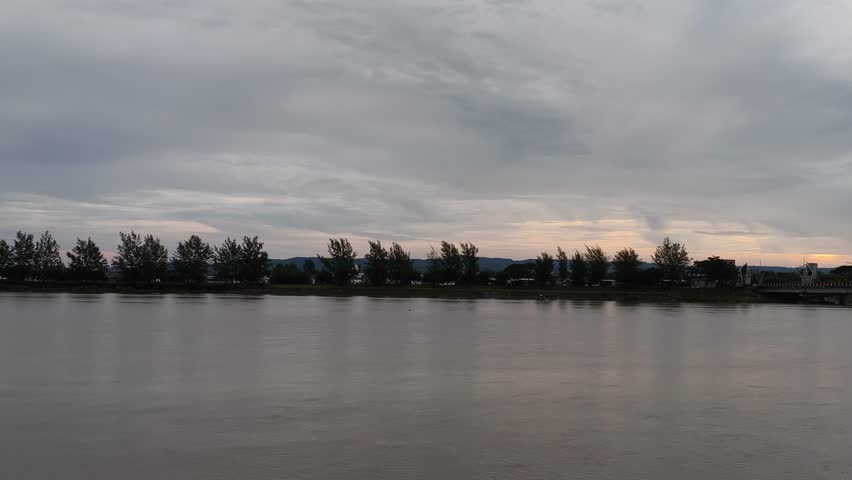 Peaceful wide river landscape at dusk with a line of trees on the far bank under a cloudy sunset sky. Calm water surface and serene nature view.