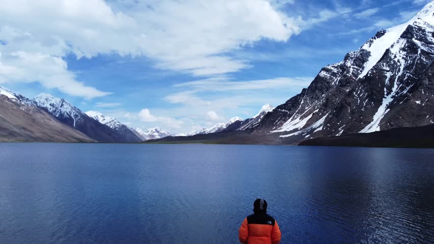 Aerial perspective of a high-altitude lake with a focus on the mountain shoreline. The drone moves along the edge of the lake, where the blue water meets the grey rock and white snow of the mountainside. The jagged edges of the peaks are clearly visible, rising steeply into the sky. This shot highlights the rugged interface between different natural elements – water, rock, and ice – in a landscape that feels ancient and untouched by human influence.