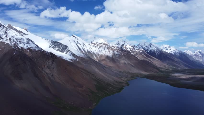 Breathtaking aerial view of a deep blue high-altitude lake nestled among snowy mountains. This drone footage descends towards a pristine alpine lake, its sapphire waters reflecting the surrounding snow-dusted peaks. The camera captures the rugged shoreline where steep mountainsides meet the water. The scene is quiet and majestic, showcasing the hidden gems found in remote mountain ranges, perfect for storytelling about environmental preservation and the sublime power of nature.