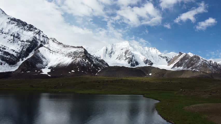 Wide-angle drone tracking shot of a vast alpine valley and distant snowy peaks. The camera moves steadily across a broad mountain pass, capturing the immense scale of the surrounding rock formations. In the background, majestic mountains rise into a clear sky, their summits cloaked in perpetual snow. The lighting captures the textures of the mountainside, from craggy outcrops to patches of hardy grass, illustrating the harsh yet beautiful environment of the high mountains.