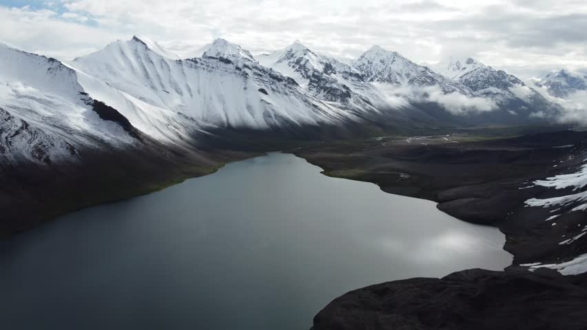 Wide landscape of a lake surrounded by snowy mountains. The drone pulls back to reveal a vast panoramic view of a mountain lake nestled in a valley of snow-covered summits. The dark water provides a stark contrast to the white peaks and the grey sky above. This footage emphasizes the scale and isolation of the region, perfect for travel features or environmental documentaries focused on the pristine beauty of high-altitude water sources and glacial landscapes.