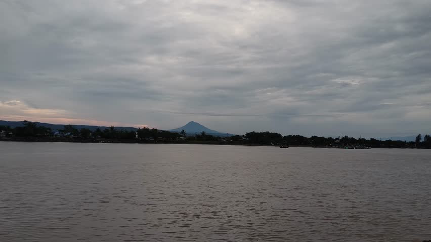 Wide tropical river landscape under a cloudy sky at sunset. Serene water view with a bridge and distant trees in a peaceful evening atmosphere.