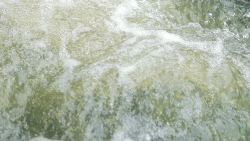 High-quality 4K resolution stock footage featuring an abstract, extreme close-up of a powerful river's surface. The camera captures the chaotic beauty of "white water" as it churns, creating a thick layer of bubbles, froth, and foam. The sunlight illuminates the water, highlighting the crystalline details of the splashes and the rapid, fluid movement of the current. This cinematic macro shot focuses entirely on the water's texture and energy, making it an ideal background or B-roll for projects involving nature's power, water resources, environmental conservation, or as a meditative visual for wellness and mindfulness content set in the tropical landscapes of Southeast Asia.