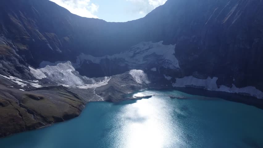 Dramatic low-angle sun over a shimmering glacial lake. The drone hovers low over the water, looking towards the sun which hangs just above the jagged horizon. The intense light creates a silver path across the lake and casts the surrounding mountains into deep relief. This cinematic shot captures the golden hour in the high mountains, highlighting the raw and powerful beauty of the natural world at the transition between day and night.