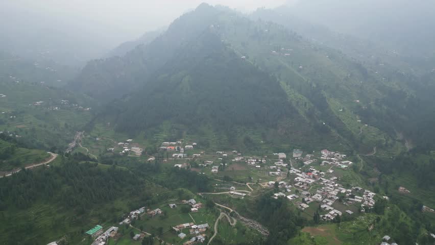High-altitude aerial view of a mountain valley with a small town. This shot captures a wide perspective of a valley floor populated with buildings, surrounded by towering green mountains. The camera drifts slowly, providing a sense of scale and showing the relationship between urban development and the vast natural landscape of the highlands.