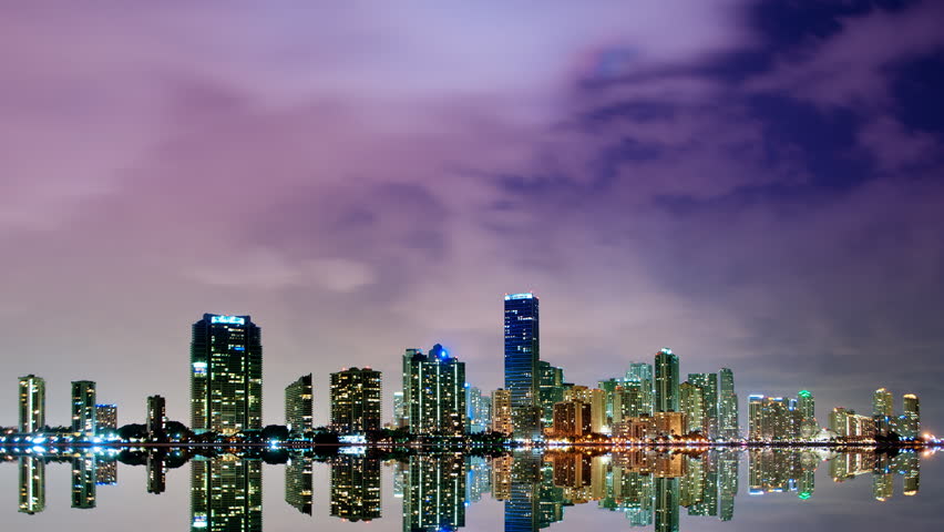 Static time lapse of the Miami skyline at night with vivid city lights, dramatic clouds moving across the sky, and a mirror-like reflection on calm water.