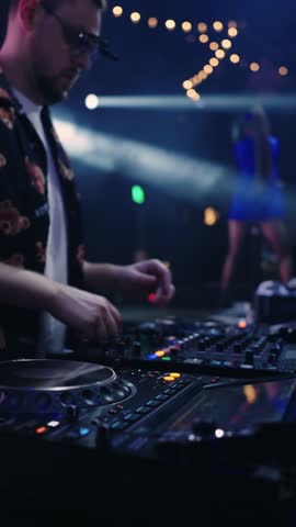 Nightclub DJ performing live electronic set on illuminated mixer console with colorful lights, energetic crowd silhouettes, and vibrant party atmosphere on dance floor