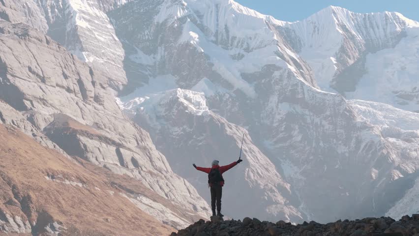 adventurer celebrates atop rocky summit, surrounded by breathtaking snow-capped mountains and clear blue sky, showcasing a moment of achievement and exploration.