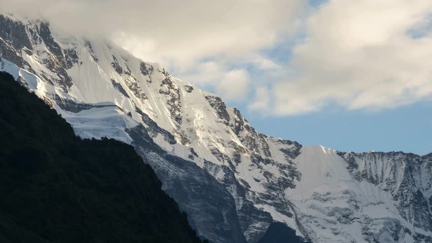 Snow-capped peaks rise against a clear blue sky, highlighting rugged mountain terrain, dramatic shadows, and vibrant cloud formations in a stunning landscape.