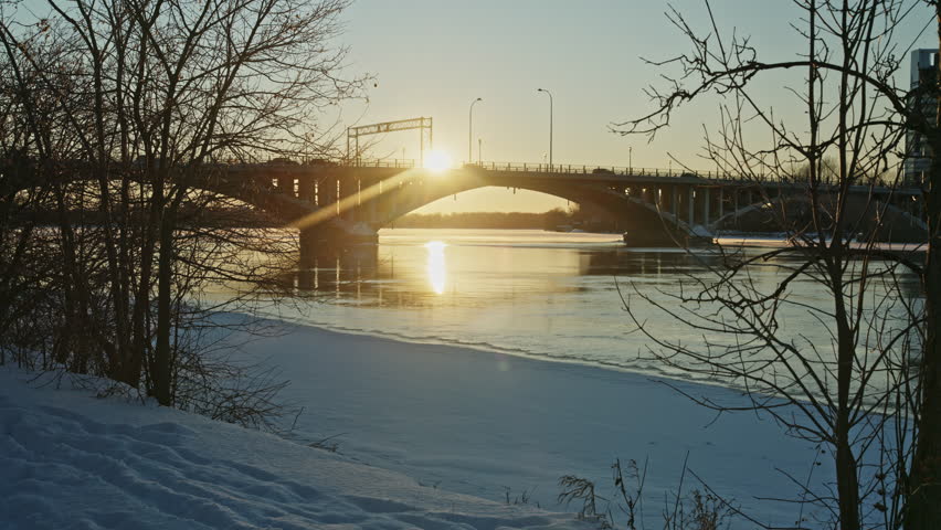Partially frozen river flowing under a bridge at winter sunset with snow banks and golden reflections. Lateral movement.