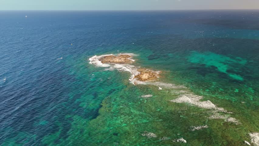 Orbital drone view from the protected Blata tal-Ghallis natural rocky island formation just emerging above the gorgeous shallow blue Mediterranean Sea, close to the Maltese shore.