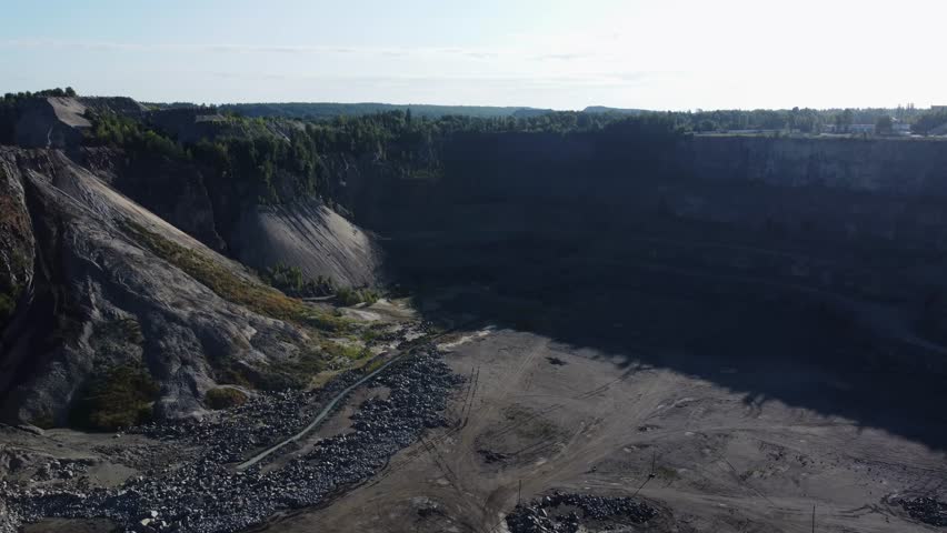 Bottom and slope of the quarry for extraction of gray granite with terraces in shadow, located among the forest, aerial view while moving forward in summer day

