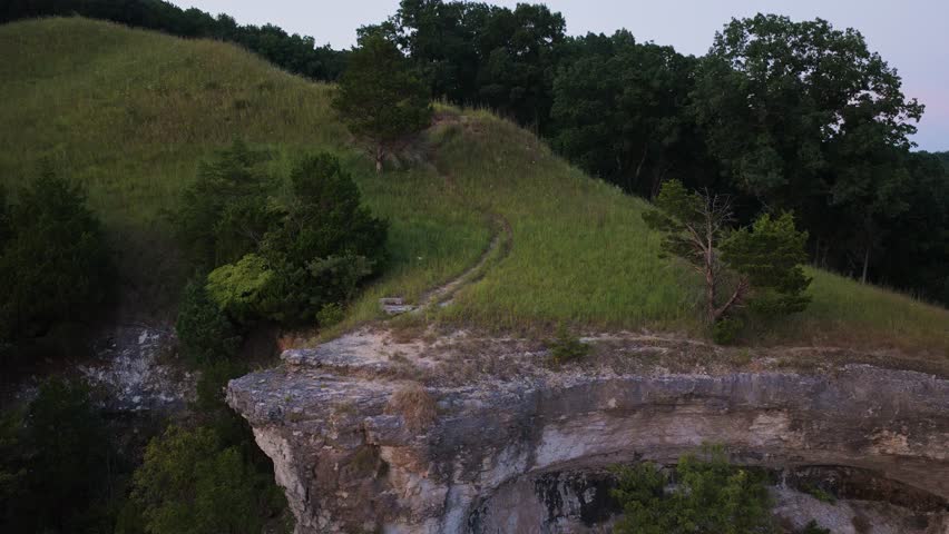 Cinematic drone footage flying toward a cliff edge and along a grassy trail at Fults Hill Prairie in Southern Illinois. The drone gains altitude to reveal the full river bluff line at sunset.