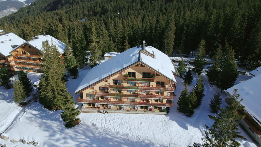 A wide aerial over a cozy wooden ski chalet in beautiful snow-covered Meribel, with skiers and snowboarders passing between pine trees on their luxury winter vacation in the French Alps.