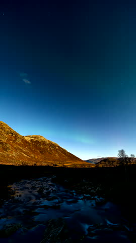 Aurora glows above a river valley and mountains at Kattfjordeidet near Tromso, Norway, vertical timelapse