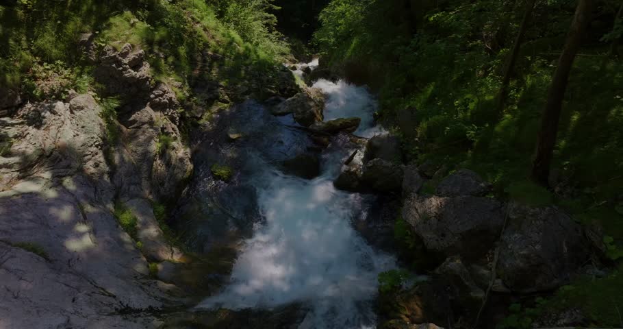 Drone tracks slowly upstream over churning blue-white water between rock walls and green forest, moving toward the hidden waterfall used as a Shamanic ceremony location.