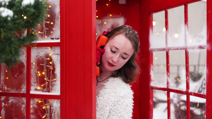 A Christmas concept, Happy woman in a fur coat posing in a red telephone booth on snowing day.