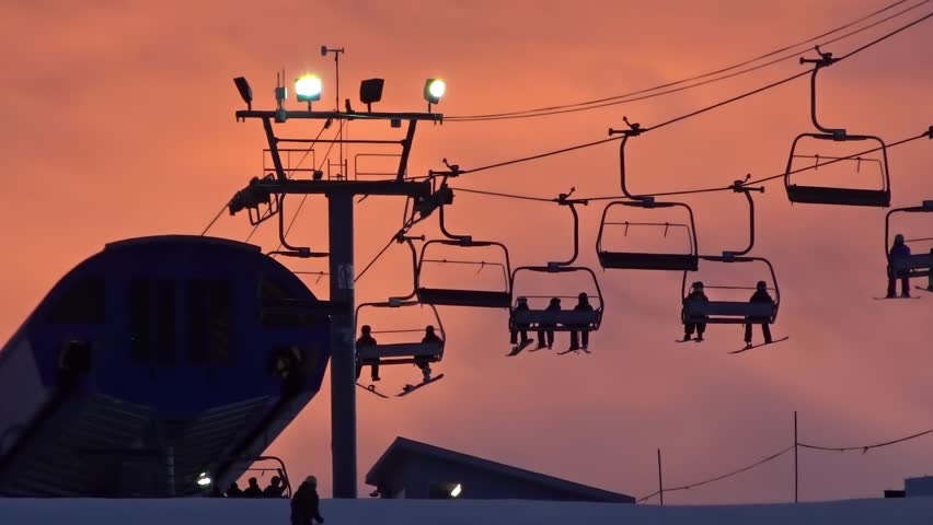 Skiers and snowboarders ride a chairlift at dusk at Calgary Olympic Park. Low-angle silhouette shot against a vibrant orange sky in Calgary, Alberta, Canada.