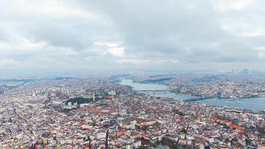 Wide aerial view of Istanbul skyline showing dense urban districts, Bosphorus waterway, bridges, and historic neighborhoods. Drone footage captures the vast cityscape under cloudy skies in Turkey. UHD