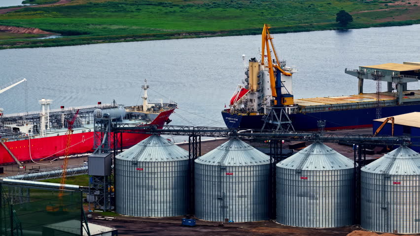 Aerial view of a riverside industrial terminal with large metallic grain silos in the foreground and cargo vessels docked, surrounded by cranes and loading structures along the waterfront.
