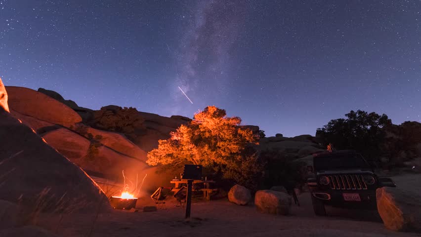 4K horizontal night time-lapse video of a campsite in Joshua Tree National Park featuring a glowing campfire and the Milky Way moving across a starry sky. The scene captures the serene desert landscape, silhouetted Joshua trees, and clear night sky with stars streaking in long-exposure motion.