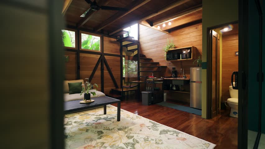 Warm and inviting interior of a rustic wooden cabin nestled in the jungle. A cozy living space with a kitchenette and a modern spiral staircase