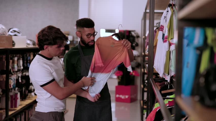 Attentive shop assistant explaining the technical features of a cycling jersey to a young male customer, helping him choose the right sportswear for his biking needs inside a specialized store