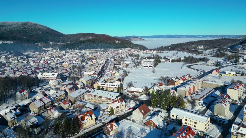 A picturesque winter scene of a snow-covered town nestled in a valley, surrounded by forests under a clear blue sky. A frozen pond awaits skaters.