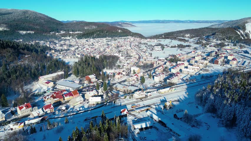 A picturesque winter scene of a snow-covered town nestled in a valley, surrounded by forests under a clear blue sky. A frozen pond awaits skaters.