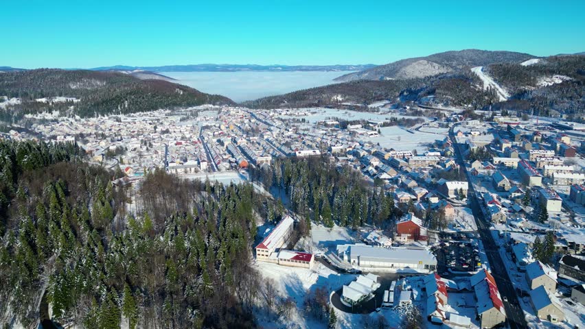 A picturesque winter scene of a snow-covered town nestled in a valley, surrounded by forests under a clear blue sky. A frozen pond awaits skaters.
